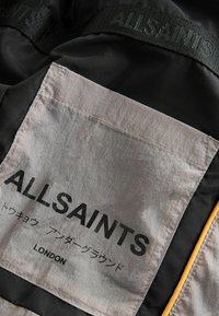 Svart tyg med en stor grå fläck där det står "ALLSAINTS" i fet svart text, accenterad med gul söm och ett texturerat band.