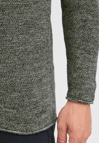 Pulover gri închis cu un model de tricot texturat, având o margine rotunjită și detalii cu manșete răsucite. Etichetă mică cu logo aproape de margine.