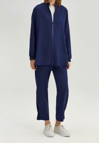 Marineblauwe zip-up jas met witte binnenkant, gecombineerd met bijpassende broek. Zachte stof, losse pasvorm en splitaccenten aan de broekspijpen. Casual schoenen.