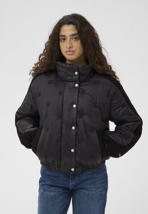 Donna con capelli lunghi e ricci che indossa una giacca imbottita nera trapuntata con bottoni a pressione e jeans blu, in piedi contro uno sfondo uniforme.