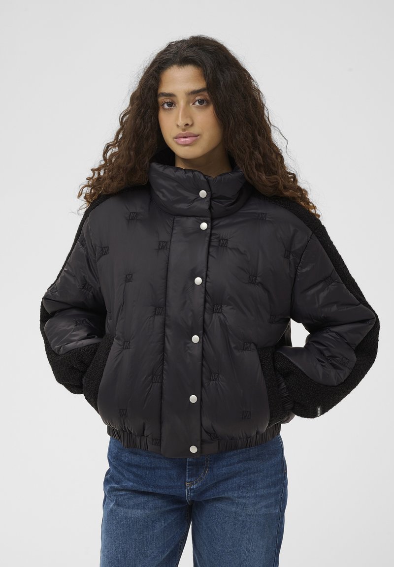 Femme aux longs cheveux bouclés portant une doudoune matelassée noire avec des boutons-pression et un jean bleu, debout devant un fond uni.