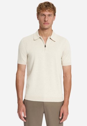 Man met lichte huid en krullend blond haar die een crème-kleurig polo met korte mouwen en een ritssluiting bij de hals draagt en een bruine broek, staand.