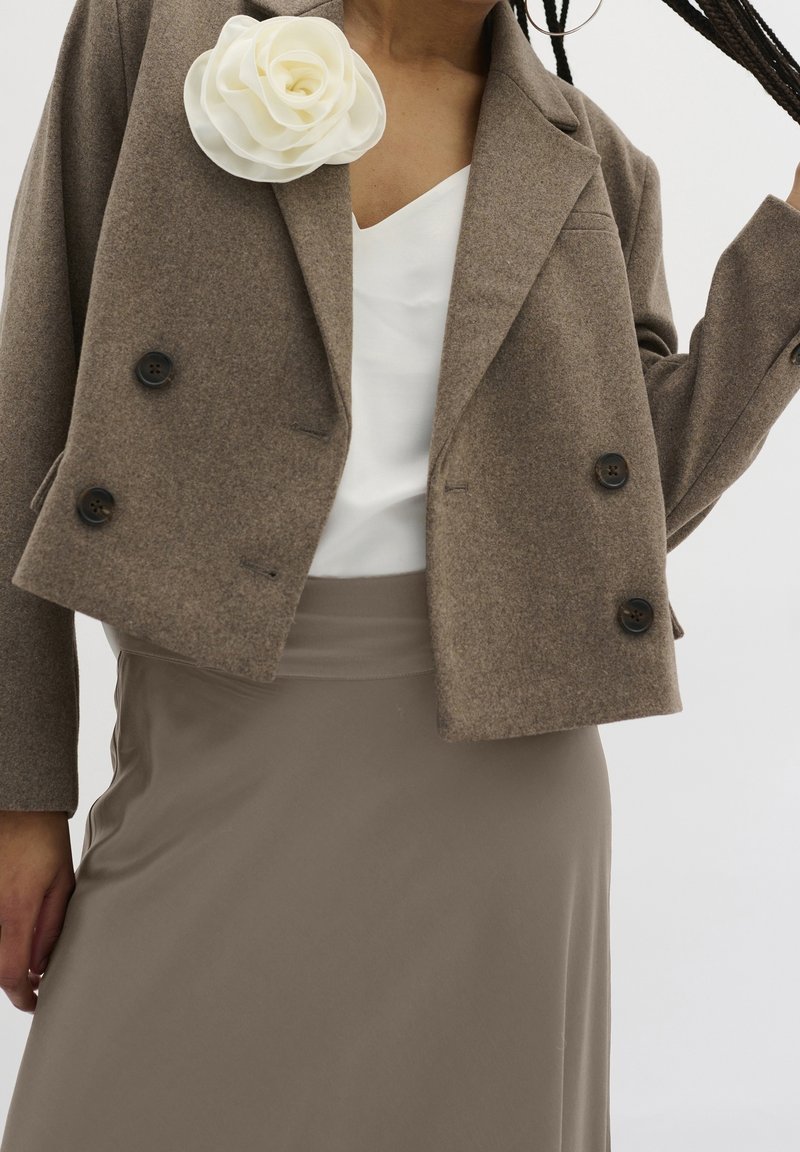 Blazer corto in misto lana marrone con doppi bottoni neri, abbinato a un top bianco a scollo a V e una gonna beige a vita alta. Spilla a forma di fiore come accento.
