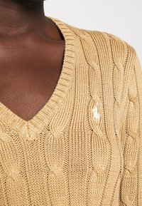 Beige neulepaita, jossa on V-pääntie, teksturoitu kabelikuvio ja pieni logotieto, sekä ribatut reunat pääntie kohdalla.