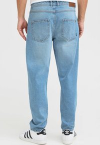 Jeans de mezclilla de color azul claro con corte relajado, que presentan una cintura alta, dos bolsillos traseros y costuras visibles. Llevados con zapatos blancos.
