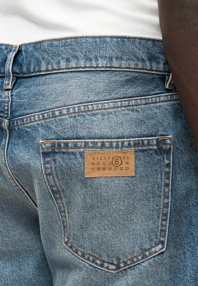 Svetlomodré rifľové nohavice sohnedou koženou náplasťou na zadnom vrecku s vytlačenými číslami. Klasický dizajn s piatimi vreckami a prešívanými detailmi.