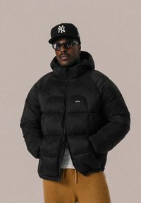 Doudoune noire avec capuche, dotée d'une fermeture éclair, de poignets côtelés et d'un petit logo. Portée par-dessus une chemise claire avec un pantalon marron texturé.