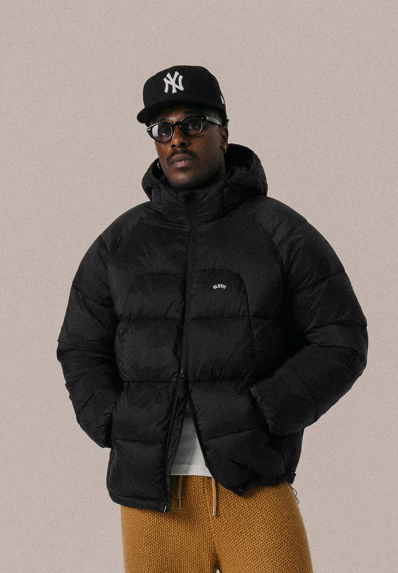 Doudoune noire avec capuche, dotée d'une fermeture éclair, de poignets côtelés et d'un petit logo. Portée par-dessus une chemise claire avec un pantalon marron texturé.