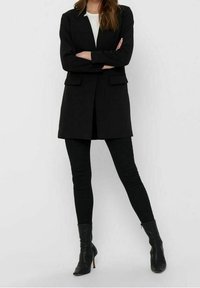Žena v černém saku ke kolenům, černých skinny kalhotách, černých kožených kotníkových botách na podpatku a bílém topu, stojící s překříženýma rukama před bílým pozadím.