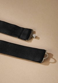 Zwarte stoffen riem met een gestructureerd patroon, voorzien van zilveren metalen sluitingen aan de uiteinden. Legt tegen een lichtbruine achtergrond.