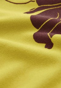 Tkanina o miękkiej, teksturowanej powierzchni w jasnym żółtym kolorze z ciemnobrązowym abstrakcyjnym wzorem, podkreślająca gładkość i żywy kontrast kolorów.