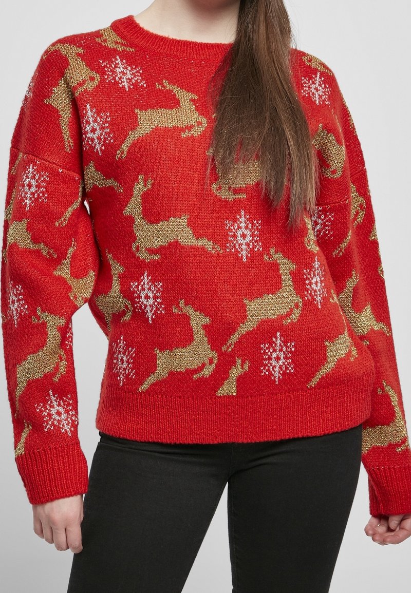 Pull rouge en tricot avec des motifs de rennes dorés et de flocons de neige blancs, porté par une personne aux longs cheveux bruns et pantalon noir.
