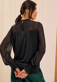 Schwarze Bluse mit langen Ärmeln, die transparente Ärmel, geraffte Bündchen und einen Ausschnitt mit Schlüsselöffnung auf der Rückseite hat, kombiniert mit grünen Hosen.