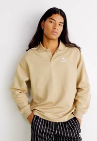 Beige Langarm-Polohemd mit Kragen und dezentem Logo, kombiniert mit schwarzen Nadelstreifen-Hosen. Glatter Stoff mit lockerer Passform.