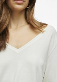 Maglione bianco a maglia con scollatura a V e bordi a coste, caratterizzato da una consistenza morbida e una vestibilità comoda. Nessun motivo o dettaglio distintivo visibile.