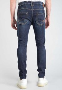 Jeans de mezclilla azul oscuro con corte slim, que cuentan con bolsillos traseros y detalles de costura clara. Completados con zapatillas blancas.