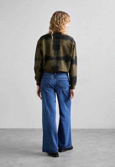 Hanorac tie-dye verde măsliniu cu accente negre, purtat împreună cu blugi drepți de denim albastru. Textură netedă și croială lejeră, include buzunare la spate.
