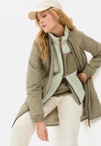 Licht olijfgroen geïsoleerd jack met een doorgestikt oppervlak en geribbelde manchetten, gedragen over een mintgroene fleece zip-up. Beige broek.