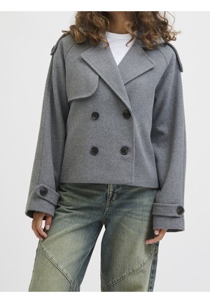 Frau trägt eine graue, doppelseitige Wolljacke mit breiten Revers und geknöpften Manschetten, kombiniert mit locker sitzenden, grünlichen Denim-Jeans.