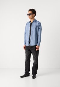 Light blue button-up shirt, black T-shirt, black jeans, and black shoes. Model wearing sunglasses, standing against a plain white background.