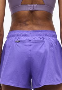 Γυναίκα που φορά μοβ αθλητικές σορτς με φερμουάρ στην πίσω τσέπη και ασορτί μοβ αθλητικό σουτιέν, εμφανιζόμενη από πίσω.