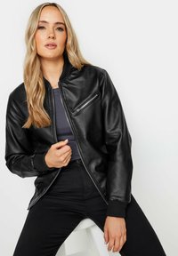 Svart läderbomberjacka med en dragkedjeficka på bröstet, ribbade ärmslut och en slät, strukturerad finish. Bärs över en grå topp.