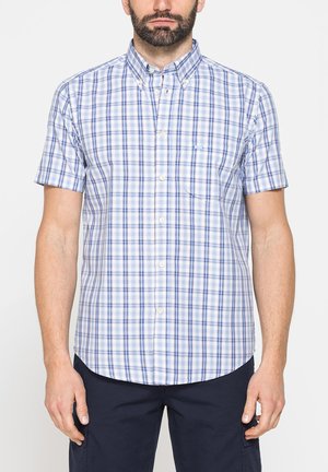 Uomo che indossa una camicia bianca a maniche corte con motivo a quadri blu e viola e pantaloni blu navy su sfondo bianco.