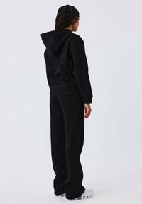 Zwarte capuchontrui met een relaxte pasvorm, gecombineerd met bijpassende zwarte sweats. De stof lijkt zacht en fleeceachtig, wat comfort biedt.