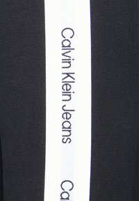 Tissu noir avec une bande verticale blanche affichant le texte "Calvin Klein Jeans". La texture est lisse avec une coupe ajustée.