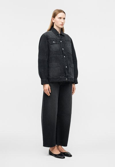 Γυναίκα που στέκεται φορώντας μαύρο, υπερμεγέθες τζιν μπουφάν με πολλές τσέπες, φαρδιά μαύρα τζιν παντελόνια και μαύρα μπαλαρίνα παπούτσια.