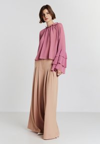 Donna che indossa una blusa rosa a maniche a volant larga abbinata a pantaloni beige a vita alta e larghi, in piedi contro uno sfondo semplice.