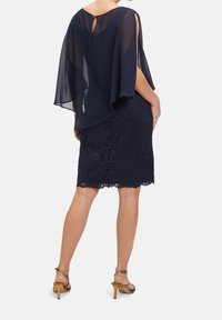 Femme portant une jupe crayon en dentelle bleu marine et un top cape transparent avec un dos ajouré, associée à des sandales à talons hauts couleur beige, debout de dos.