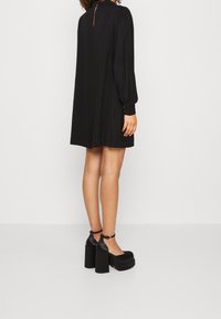 Abito lungo nero a maniche lunghe con una silhouette morbida, caratterizzato da un collo alto e un dettaglio a chiave sul retro. Abbinato a tacchi a piattaforma.