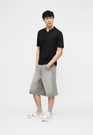 Jaunas vyras stovintis su rankomis kišenėse, dėvintis juodą trumpomis rankovėmis marškinėlį, pilkus kelio ilgio šortus, pilkas kojines ir baltus sportinius batelius.