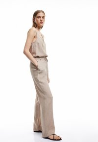 Mono de lino beige con piernas anchas, escote en forma de v y cinturón anudado en la cintura. Textura suave y diseño minimalista.