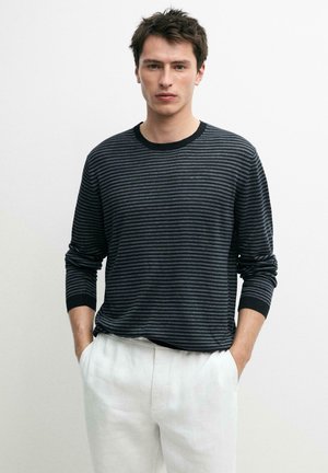 Jeune homme aux cheveux courts et foncés portant un t-shirt à manches longues rayé noir et gris et un pantalon blanc, mains dans les poches, fond neutre.