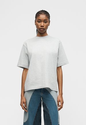 Ung kvinde med flettet hår iført en løs lysegrå kortærmet skjorte og blå patchwork-jeans, stående mod en ensfarvet baggrund.