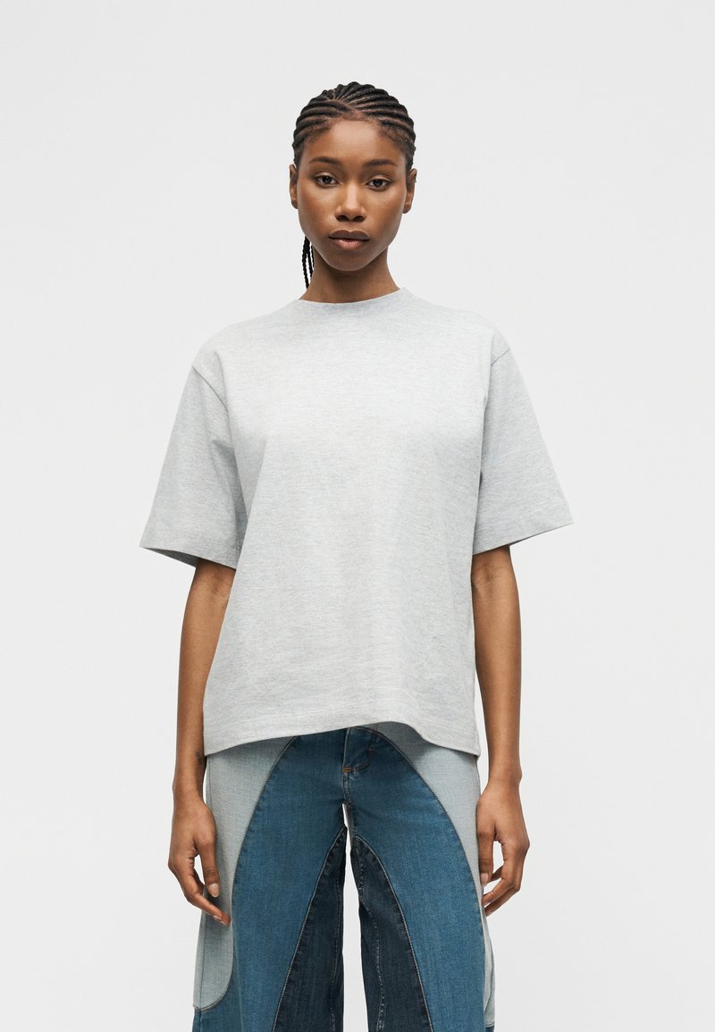 Νεαρή γυναίκα με πλεκτά μαλλιά, που φορά χαλαρό ανοιχτό γκρι κοντομάνικο πουκάμισο και τζιν με σχέδια μπλε, να στέκεται μπροστά σε απλό φόντο.