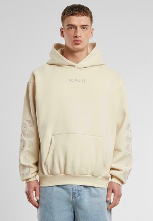 Sweat à capuche crème avec poche frontale, poignets côtelés et accents à motif paisley sur les deux manches. Logo "KANI" brodé sur la poitrine.