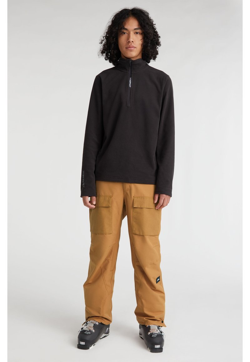 Schwarzer Fleece-Pullover mit hohem Kragen und Reißverschluss, kombiniert mit hellbraunen Cargo-Hosen mit großen Taschen, getragen mit schwarzen Skischuhen.