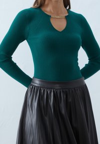 Femme portant un pull côtelé turquoise avec un détail chaîne dorée au niveau de l'encolure et une jupe en cuir plissée noire.