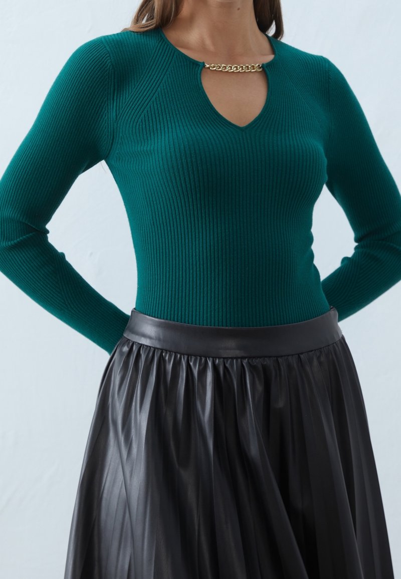 Femme portant un pull côtelé turquoise avec un détail chaîne dorée au niveau de l'encolure et une jupe en cuir plissée noire.