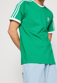 Grön bomullströja med vit ribbad rund halsringning och ärmar, med tre vita ränder på axlarna och en liten logotyp på bröstet.