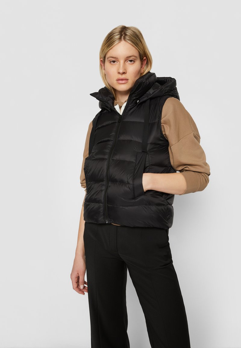 Czarna kamizelka puchowa z kapturem, z pikowanym wzorem, z zamkiem z przodu i bocznymi kieszeniami. Nosi się ją na jasnobrązowej bluzce i czarnych spodniach.