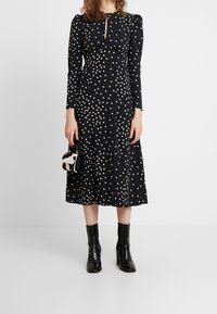 Svart midiklänning med vita prickar, långa ärmar och nyckelhålsurringning. Kombination med svarta klackstövlar och en liten handväska.