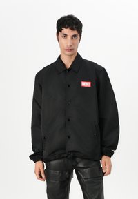 Svart vindjakke med knappestolpe foran, elastiske mansjetter, og snor i kanten. Har et rødt "DIESEL" logo-lapp på brystet.
