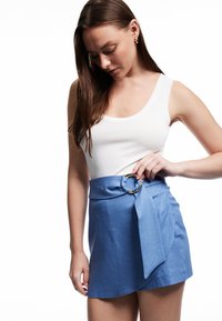 Jonge vrouw die een gouden ringriem aanpast op een blauwe wikkelrok, met een witte tanktop en gouden creolen tegen een witte achtergrond.