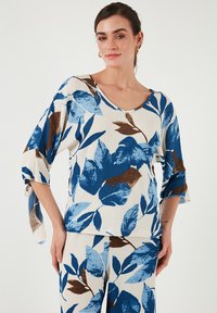 Top in maglia floreale con motivi a foglia blu e marrone su sfondo crema, con scollatura ampia e maniche a tre quarti.