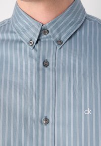 Chemise bleu clair à boutons avec rayures blanches verticales, col boutonné et petit logo blanc "ck" sur la poitrine.