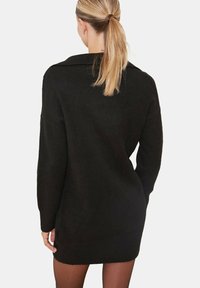 Robe pull noire en maille avec col, manches longues et coupe décontractée. La texture est douce, et le vêtement arrive à mi-cuisse.